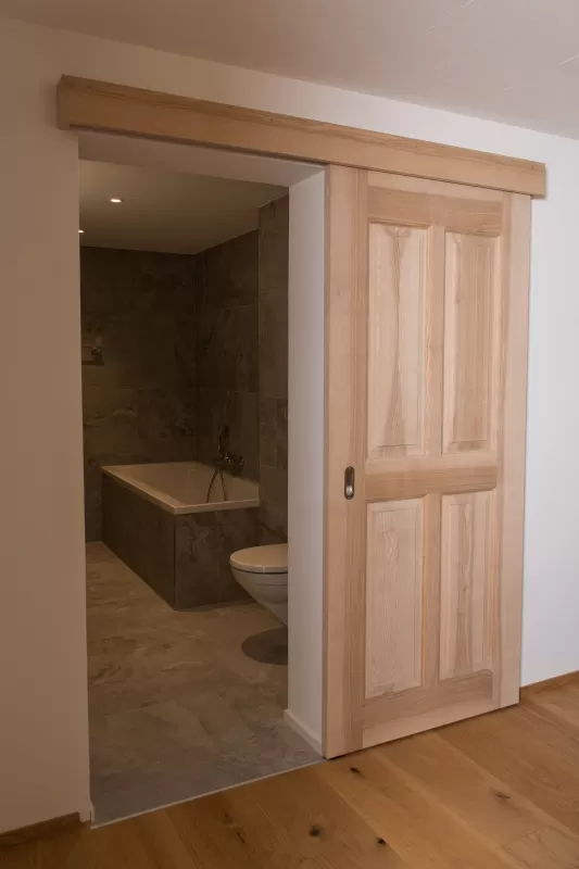 Massgefertigte Schiebetür aus Schweizer Eschenholz in heller, natürlicher Optik. Die Tür öffnet den Blick in ein modern gestaltetes Badezimmer mit grossformatigen Steinplatten, einer integrierten Badewanne und einer zeitlosen, schlichten Ausstattung. Die Kombination aus warmem Holz und reduzierten Materialien schafft eine ruhige, hochwertige Wohnatmosphäre.