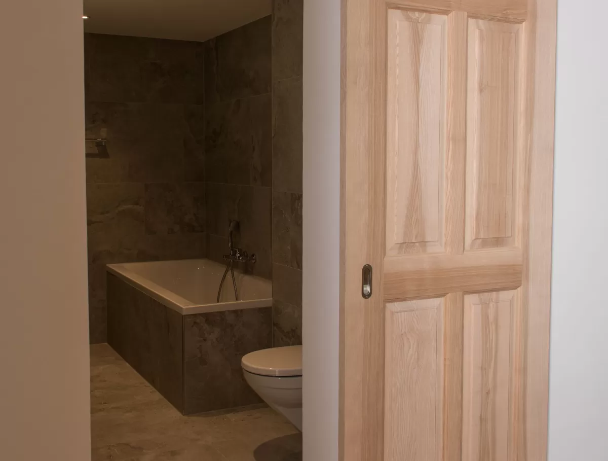 Massgefertigte Schiebetür aus Schweizer Eschenholz in heller, natürlicher Optik. Die Tür öffnet den Blick in ein modern gestaltetes Badezimmer mit grossformatigen Steinplatten, einer integrierten Badewanne und einer zeitlosen, schlichten Ausstattung. Die Kombination aus warmem Holz und reduzierten Materialien schafft eine ruhige, hochwertige Wohnatmosphäre.
