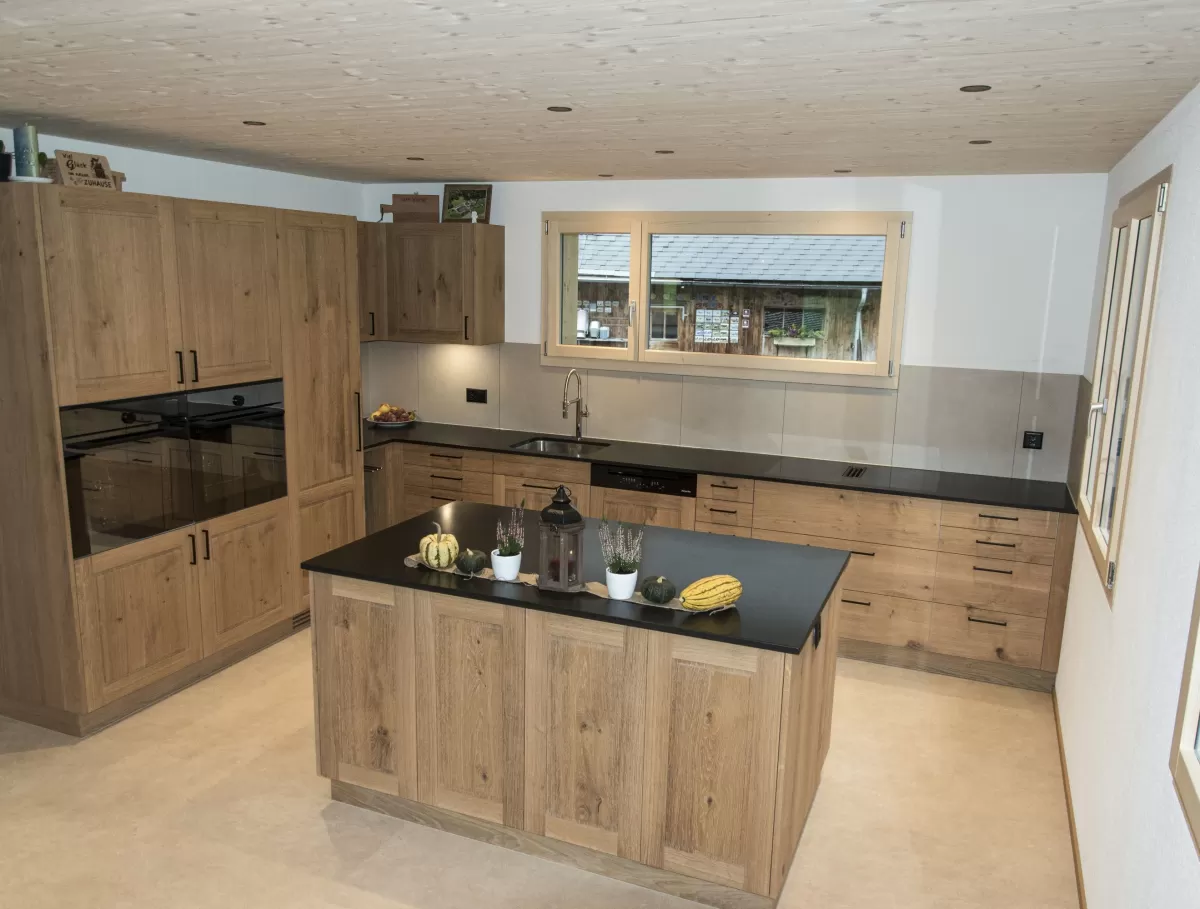 Massgefertigte Küche aus warmem Eichenholz mit zentraler Kochinsel und dunkler Arbeitsplatte. Die Kombination aus natürlichen Holzfronten, klaren Linien und modernen Einbaugeräten schafft eine zeitlose und funktionale Raumgestaltung. Grosszügige Fenster sorgen für viel Tageslicht und unterstreichen die helle, freundliche Wohnatmosphäre.