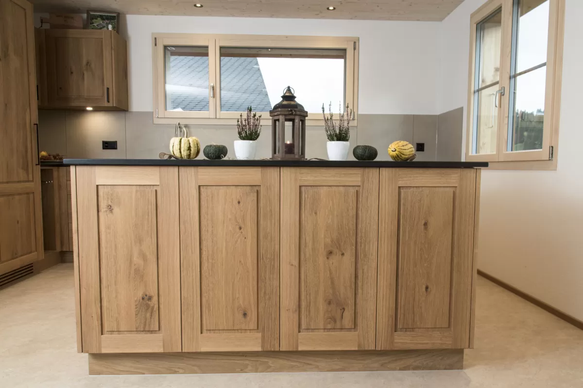 Massgefertigte Küche aus warmem Eichenholz mit zentraler Kochinsel und dunkler Arbeitsplatte. Die Kombination aus natürlichen Holzfronten, klaren Linien und modernen Einbaugeräten schafft eine zeitlose und funktionale Raumgestaltung. Grosszügige Fenster sorgen für viel Tageslicht und unterstreichen die helle, freundliche Wohnatmosphäre.