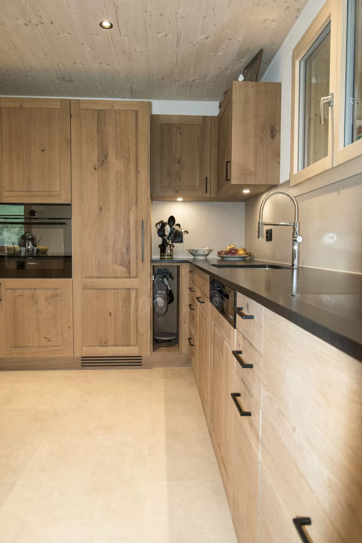 Massgefertigte Küche aus warmem Eichenholz mit zentraler Kochinsel und dunkler Arbeitsplatte. Die Kombination aus natürlichen Holzfronten, klaren Linien und modernen Einbaugeräten schafft eine zeitlose und funktionale Raumgestaltung. Grosszügige Fenster sorgen für viel Tageslicht und unterstreichen die helle, freundliche Wohnatmosphäre.