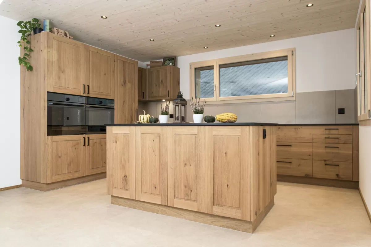 Massgefertigte Küche aus warmem Eichenholz mit zentraler Kochinsel und dunkler Arbeitsplatte. Die Kombination aus natürlichen Holzfronten, klaren Linien und modernen Einbaugeräten schafft eine zeitlose und funktionale Raumgestaltung. Grosszügige Fenster sorgen für viel Tageslicht und unterstreichen die helle, freundliche Wohnatmosphäre.
