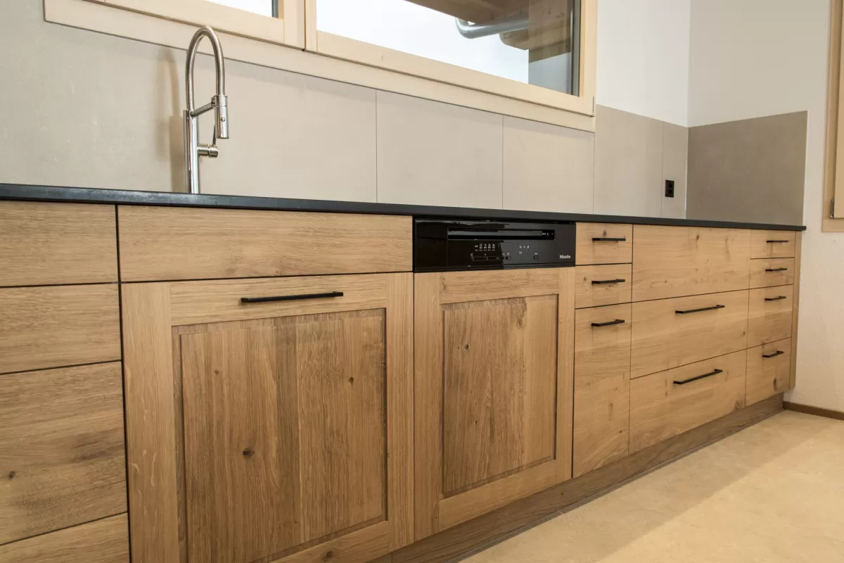 Massgefertigte Küche aus warmem Eichenholz mit zentraler Kochinsel und dunkler Arbeitsplatte. Die Kombination aus natürlichen Holzfronten, klaren Linien und modernen Einbaugeräten schafft eine zeitlose und funktionale Raumgestaltung. Grosszügige Fenster sorgen für viel Tageslicht und unterstreichen die helle, freundliche Wohnatmosphäre.