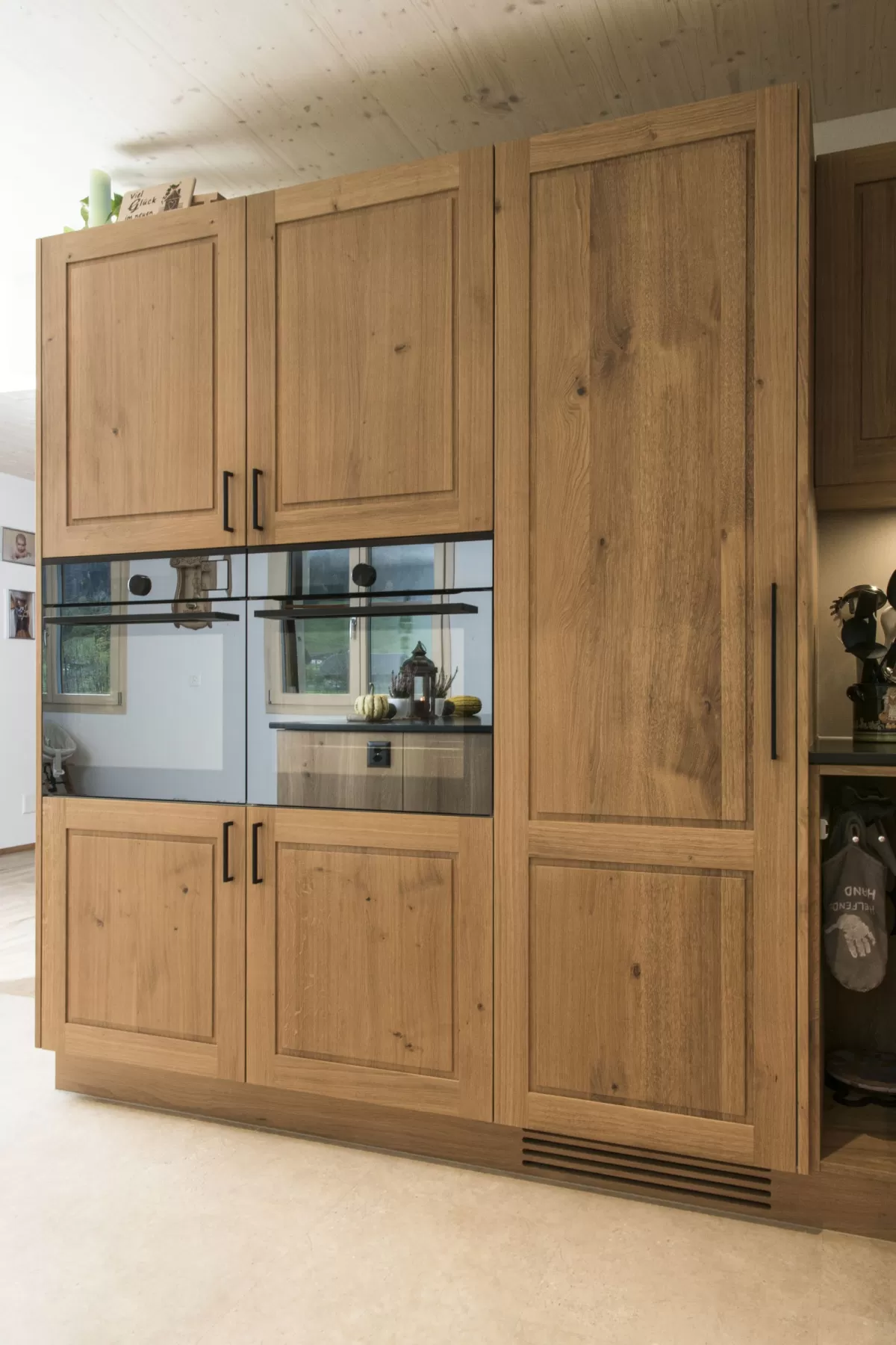 Massgefertigte Küche aus warmem Eichenholz mit zentraler Kochinsel und dunkler Arbeitsplatte. Die Kombination aus natürlichen Holzfronten, klaren Linien und modernen Einbaugeräten schafft eine zeitlose und funktionale Raumgestaltung. Grosszügige Fenster sorgen für viel Tageslicht und unterstreichen die helle, freundliche Wohnatmosphäre.