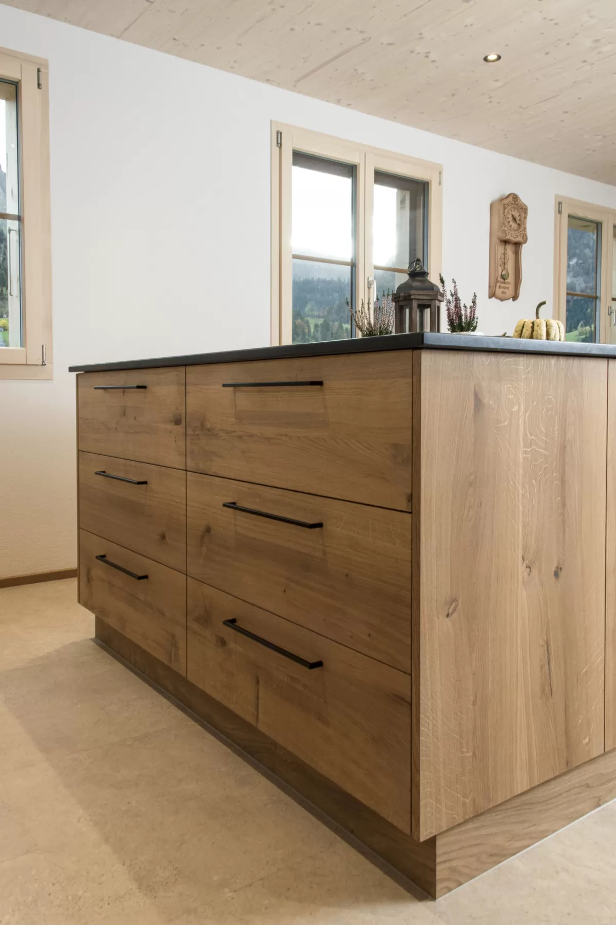 Massgefertigte Küche aus warmem Eichenholz mit zentraler Kochinsel und dunkler Arbeitsplatte. Die Kombination aus natürlichen Holzfronten, klaren Linien und modernen Einbaugeräten schafft eine zeitlose und funktionale Raumgestaltung. Grosszügige Fenster sorgen für viel Tageslicht und unterstreichen die helle, freundliche Wohnatmosphäre.