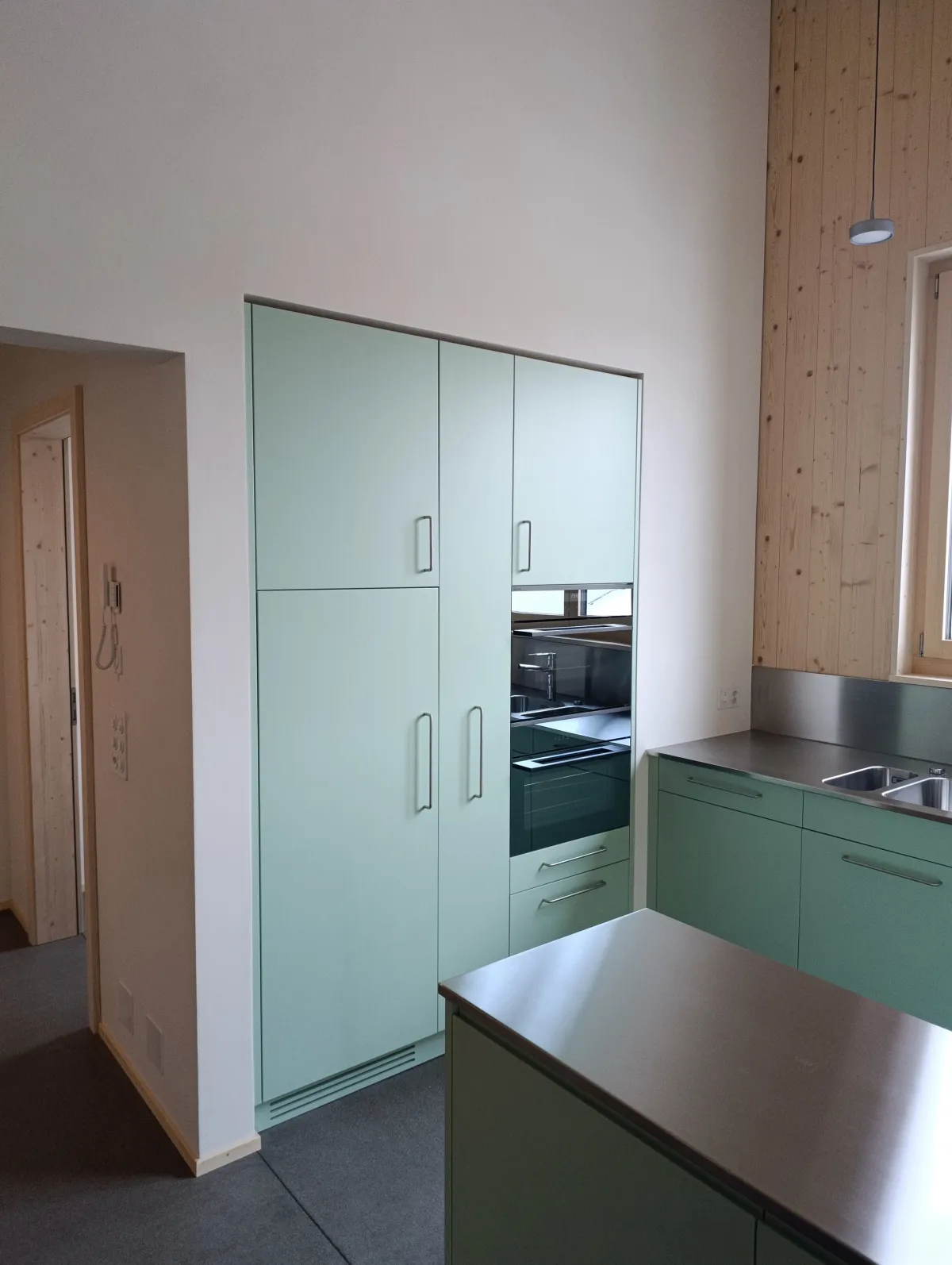 Moderne offene Küche mit rosafarbenen Fronten, Kochinsel und Arbeitsflächen aus Edelstahl. Die helle Holzdecke und die mit Holz verkleidete Fensterwand setzen einen warmen Kontrast zum dunklen Boden und zur modernen Kücheneinrichtung.