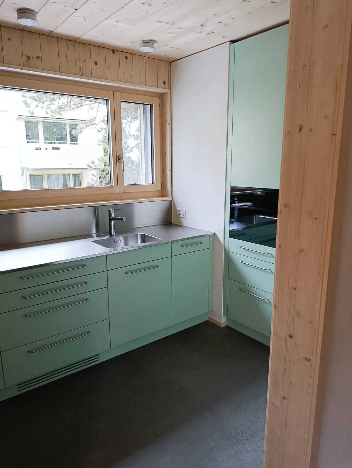 Moderne offene Küche mit rosafarbenen Fronten, Kochinsel und Arbeitsflächen aus Edelstahl. Die helle Holzdecke und die mit Holz verkleidete Fensterwand setzen einen warmen Kontrast zum dunklen Boden und zur modernen Kücheneinrichtung.