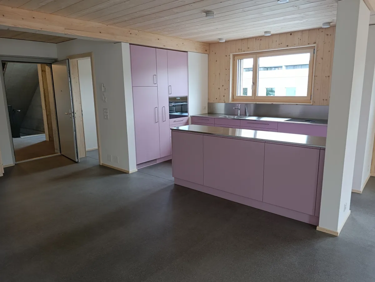 Moderne offene Küche mit rosafarbenen Fronten, Kochinsel und Arbeitsflächen aus Edelstahl. Die helle Holzdecke und die mit Holz verkleidete Fensterwand setzen einen warmen Kontrast zum dunklen Boden und zur modernen Kücheneinrichtung.