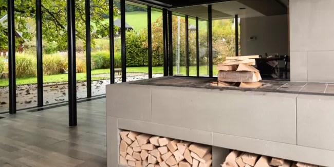 Moderner Essbereich mit grosser Glasfront und Blick in den Garten. In der Mitte steht ein runder Holztisch mit schwarzen Stühlen auf einem hellen Holzboden. Das natürliche Licht schafft eine offene, freundliche Atmosphäre.