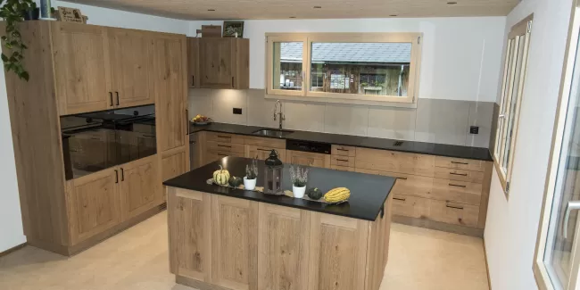 Massgefertigte Küche aus warmem Eichenholz mit zentraler Kochinsel und dunkler Arbeitsplatte. Die Kombination aus natürlichen Holzfronten, klaren Linien und modernen Einbaugeräten schafft eine zeitlose und funktionale Raumgestaltung. Grosszügige Fenster sorgen für viel Tageslicht und unterstreichen die helle, freundliche Wohnatmosphäre.