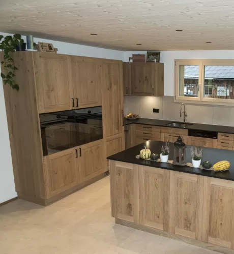 Massgefertigte Küche aus warmem Eichenholz mit zentraler Kochinsel und dunkler Arbeitsplatte. Die Kombination aus natürlichen Holzfronten, klaren Linien und modernen Einbaugeräten schafft eine zeitlose und funktionale Raumgestaltung. Grosszügige Fenster sorgen für viel Tageslicht und unterstreichen die helle, freundliche Wohnatmosphäre.
