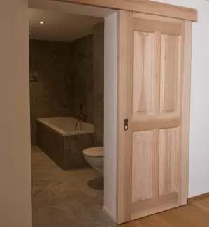 Massgefertigte Schiebetür aus Schweizer Eschenholz in heller, natürlicher Optik. Die Tür öffnet den Blick in ein modern gestaltetes Badezimmer mit grossformatigen Steinplatten, einer integrierten Badewanne und einer zeitlosen, schlichten Ausstattung. Die Kombination aus warmem Holz und reduzierten Materialien schafft eine ruhige, hochwertige Wohnatmosphäre.