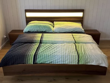 Ein modernes Doppelbett aus dunkel gemasertem Holz steht im Mittelpunkt des Schlafzimmers. Das Kopfteil ist mit einem horizontalen, warm leuchtenden Lichtstreifen versehen, der den Raum sanft ausleuchtet. Auf dem Bett liegt eine Bettwäsche in harmonischen Grün-, Blau- und Weißtönen mit einem feinen Linienmuster. Links und rechts befinden sich passende Nachttische aus dem gleichen Holz. Der Raum hat einen hellen Holzboden sowie weiss vertäfelte Wände, die eine ruhige, natürliche Atmosphäre schaffen.