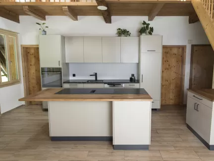 Moderne Küche mit heller Einbauküche, Kochinsel mit Holzarbeitsplatte und integrierten Geräten in einem offenen Wohnraum mit sichtbaren Holzbalken.