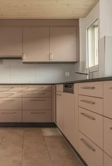 Moderne Küche mit matten, roséfarbenen Fronten, Edelstahlgriffen und hellbraunem Fliesenboden. Die Küche hat eine L-Form, eine dunkle Arbeitsplatte und eine Holzdecke mit natürlicher Maserung.