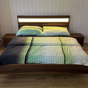 Ein modernes Doppelbett aus dunkel gemasertem Holz steht im Mittelpunkt des Schlafzimmers. Das Kopfteil ist mit einem horizontalen, warm leuchtenden Lichtstreifen versehen, der den Raum sanft ausleuchtet. Auf dem Bett liegt eine Bettwäsche in harmonischen Grün-, Blau- und Weißtönen mit einem feinen Linienmuster. Links und rechts befinden sich passende Nachttische aus dem gleichen Holz. Der Raum hat einen hellen Holzboden sowie weiss vertäfelte Wände, die eine ruhige, natürliche Atmosphäre schaffen.
