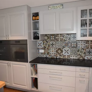 Individuell gestaltete Küche mit hellen Fronten, integrierten Einbaugeräten und einer grosszügigen Kochinsel mit massiver Holzarbeitsplatte. Der gemusterte Fliesenspiegel setzt einen dekorativen Akzent und bildet einen spannenden Kontrast zu den klaren Linien der Möblierung. Warme Holzböden und grosse Fensterflächen sorgen für eine einladende, helle Wohnatmosphäre mit Blick ins Grüne