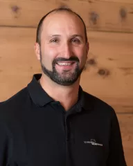 Porträt eines lächelnden Mannes mit kurzem Haar und Bart in einem schwarzen Polohemd mit dem Logo der Schreinerei Ellenberger, vor einer Holz-Wand im Hintergrund.