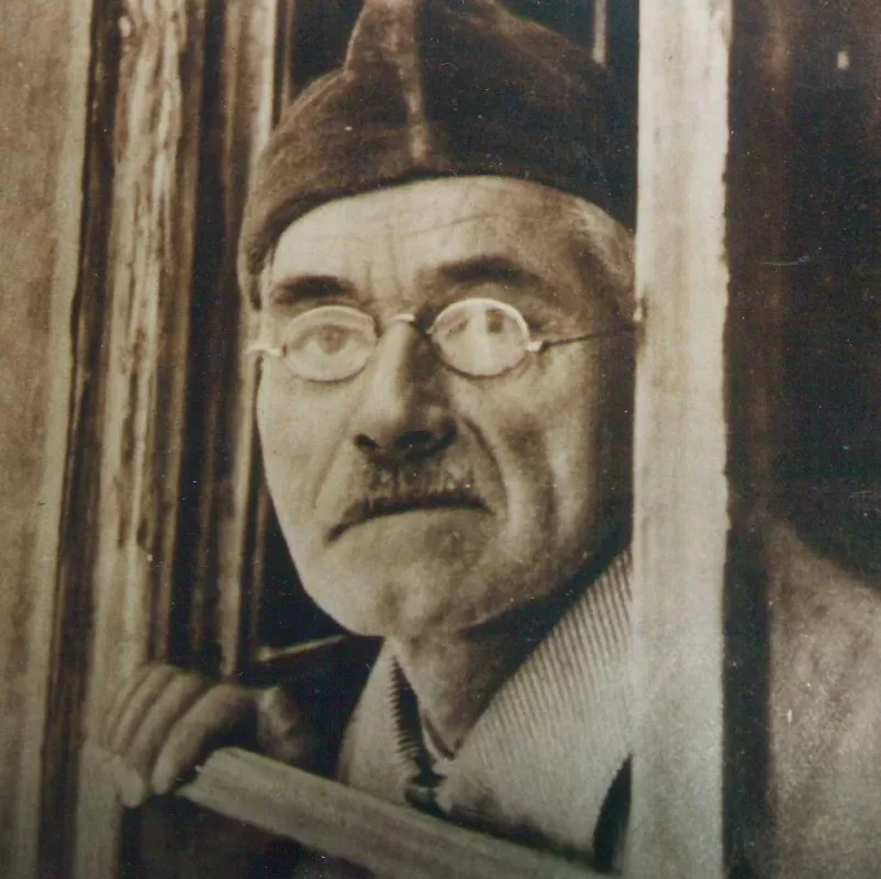 Historische Schwarz-Weiss-Aufnahme eines älteren Mannes mit Brille, Mütze und Schnurrbart, der aus einem Fenster schaut. Sein Gesichtsausdruck wirkt nachdenklich und ruhig.