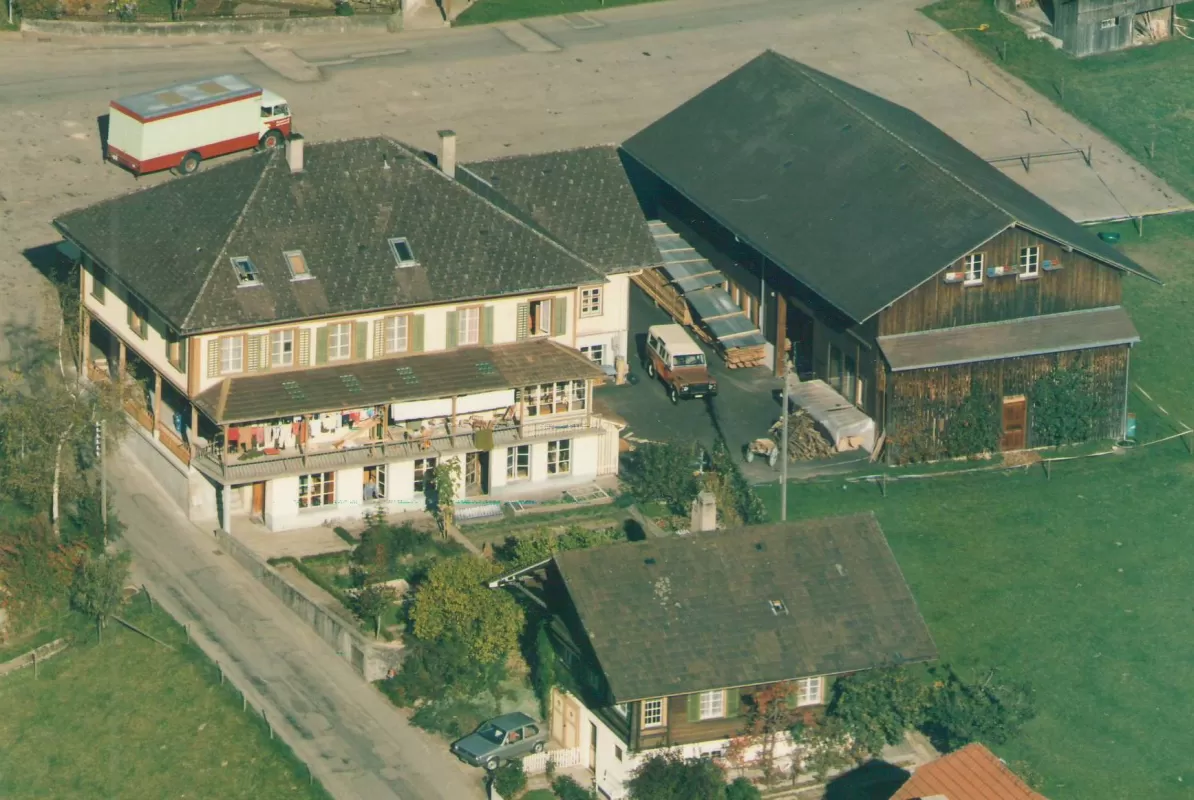 Historische Luftaufnahme der Schreinerei Eichenberger in Trub. Zu sehen sind das Hauptwohnhaus mit Balkon, die Werkstatt aus Holz sowie ein Innenhof mit parkierten Fahrzeugen und umliegendem Grünland. Im Hintergrund steht ein Lieferwagen auf dem Platz.