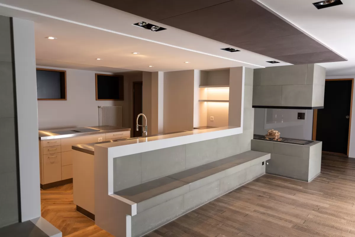 Moderne, offen gestaltete Küche mit klaren Linien und hochwertiger Verarbeitung. Sichtbar sind eine Kochinsel mit Edelstahloberfläche, integrierter Spüle und Sitzbank aus Steinoptik sowie ein offener Kamin im Hintergrund. Holzfussboden und dezente Beleuchtung schaffen eine warme Atmosphäre.