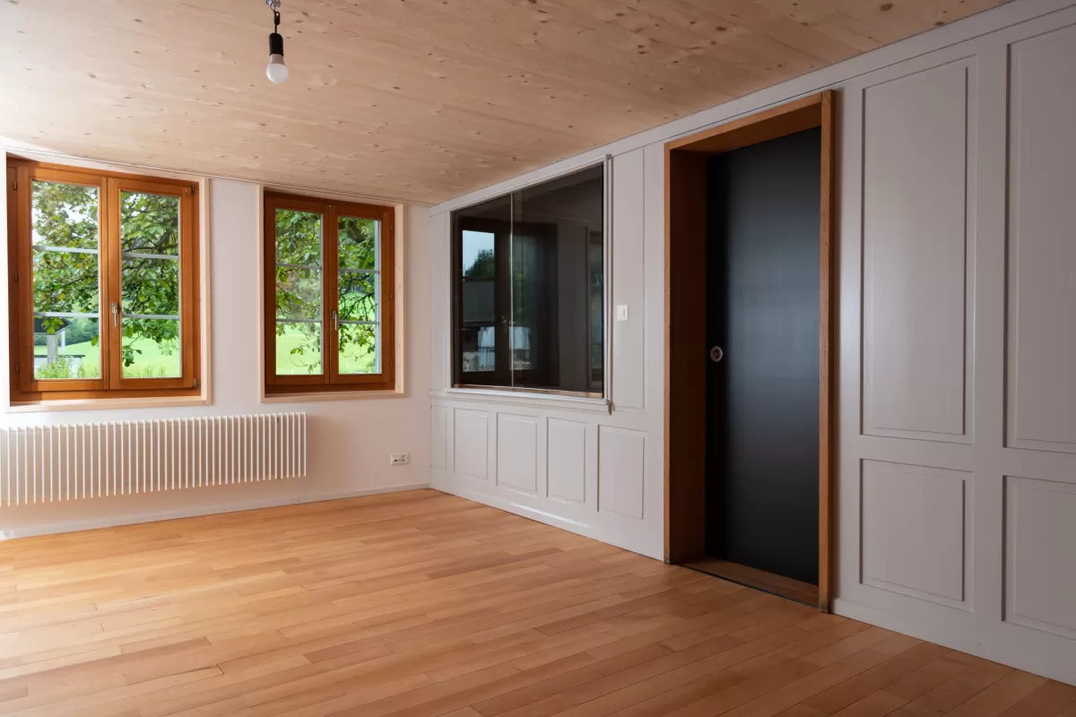 Helles, modern renoviertes Zimmer mit Holzdecke, Parkettboden und grossen Fenstern mit Holzrahmen. An der Wand befinden sich eingebaute Schrankelemente und eine schwarze Schiebetür. Der Raum wirkt natürlich, warm und klar gestaltet.