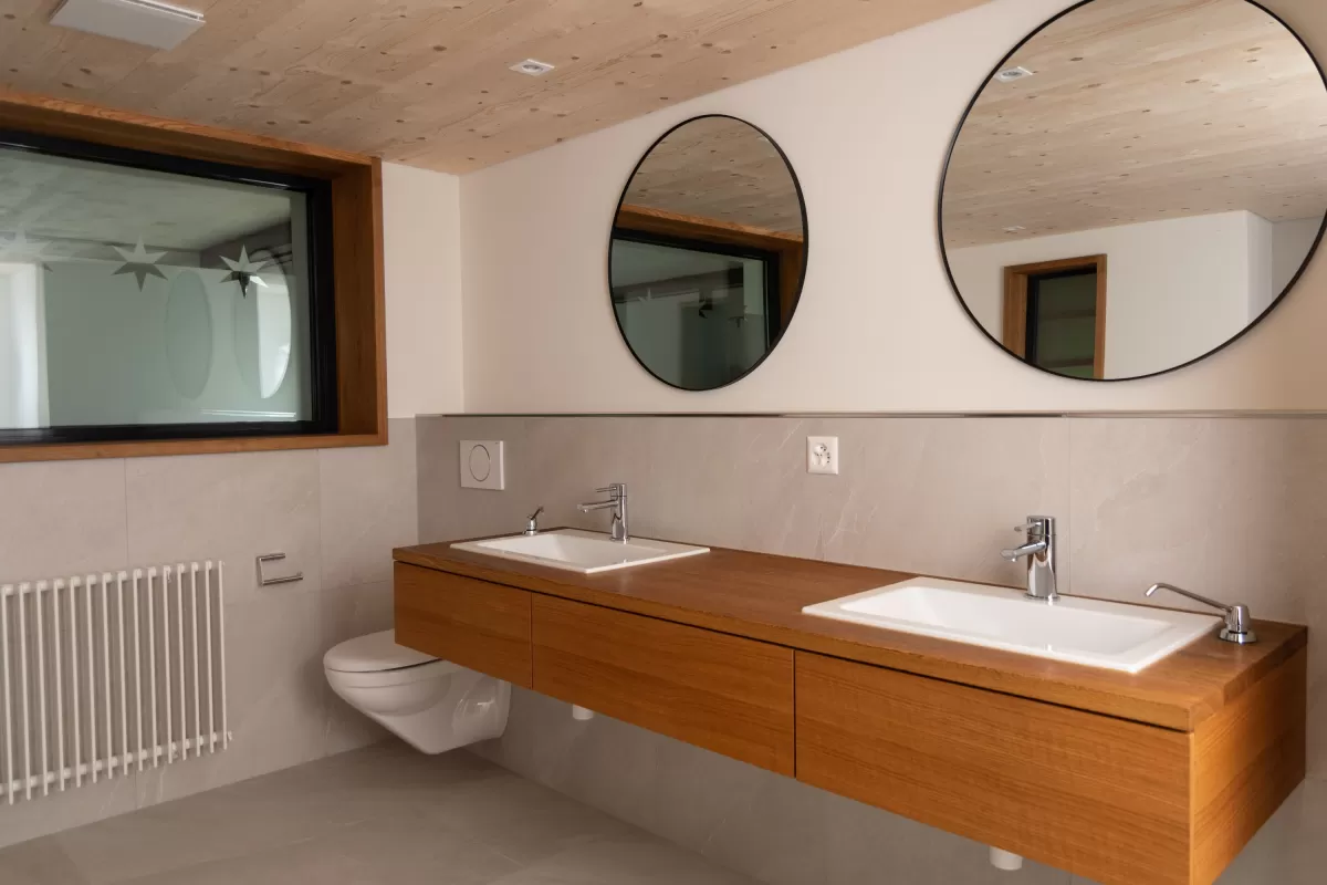 Moderner Wohnraum mit offenem Kamin und klarer, minimalistischer Architektur. Der Raum kombiniert helle Flächen mit grauen Stein- und Holzelementen, integriertem Sitzbereich und einer offenen Küche mit Edelstahlarmatur. Dezente Beleuchtung und Holzboden sorgen für eine warme Atmosphäre.