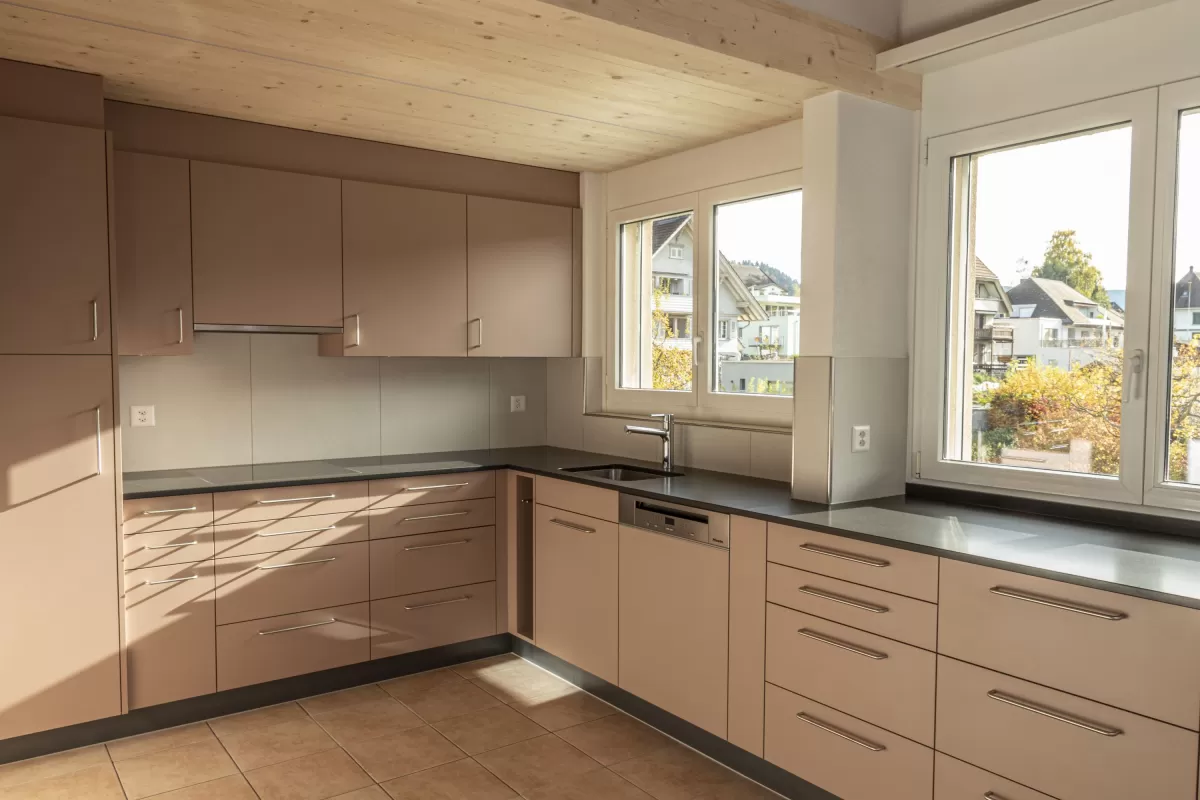 Moderne Küche mit matten, roséfarbenen Fronten, Edelstahlgriffen und hellbraunem Fliesenboden. Die Küche hat eine L-Form, eine dunkle Arbeitsplatte und eine Holzdecke mit natürlicher Maserung.