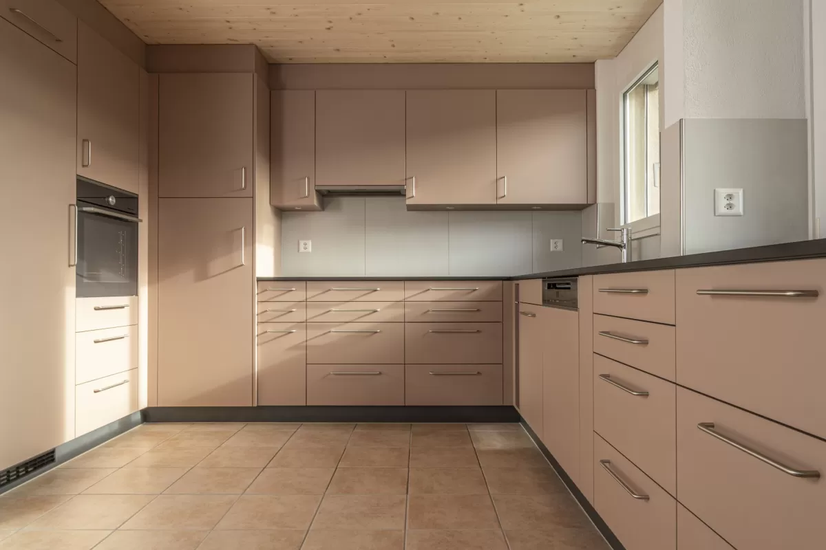 Moderne Küche mit matten, roséfarbenen Fronten, Edelstahlgriffen und hellbraunem Fliesenboden. Die Küche hat eine L-Form, eine dunkle Arbeitsplatte und eine Holzdecke mit natürlicher Maserung.