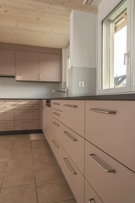 Moderne Küche mit matten, roséfarbenen Fronten, Edelstahlgriffen und hellbraunem Fliesenboden. Die Küche hat eine L-Form, eine dunkle Arbeitsplatte und eine Holzdecke mit natürlicher Maserung.