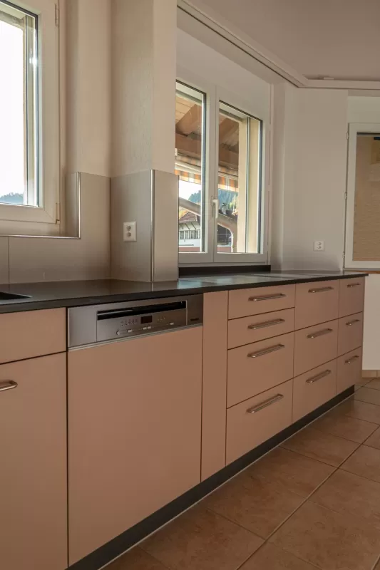 Moderne Küche mit matten, roséfarbenen Fronten, Edelstahlgriffen und hellbraunem Fliesenboden. Die Küche hat eine L-Form, eine dunkle Arbeitsplatte und eine Holzdecke mit natürlicher Maserung.