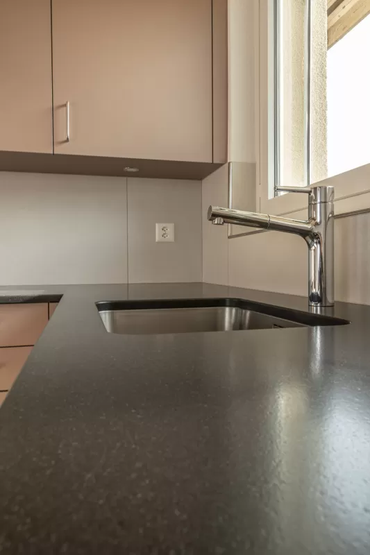 Moderne Küche mit matten, roséfarbenen Fronten, Edelstahlgriffen und hellbraunem Fliesenboden. Die Küche hat eine L-Form, eine dunkle Arbeitsplatte und eine Holzdecke mit natürlicher Maserung.