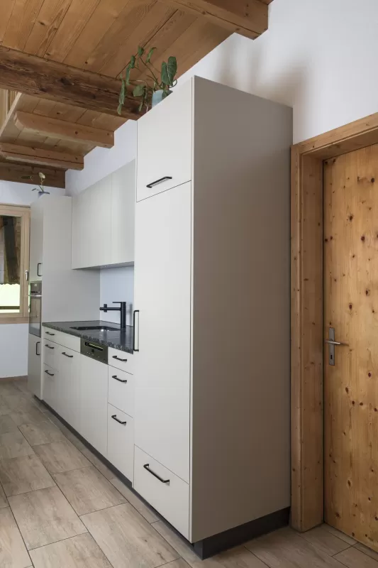 Moderne Küche mit heller Einbauküche, Kochinsel mit Holzarbeitsplatte und integrierten Geräten in einem offenen Wohnraum mit sichtbaren Holzbalken.