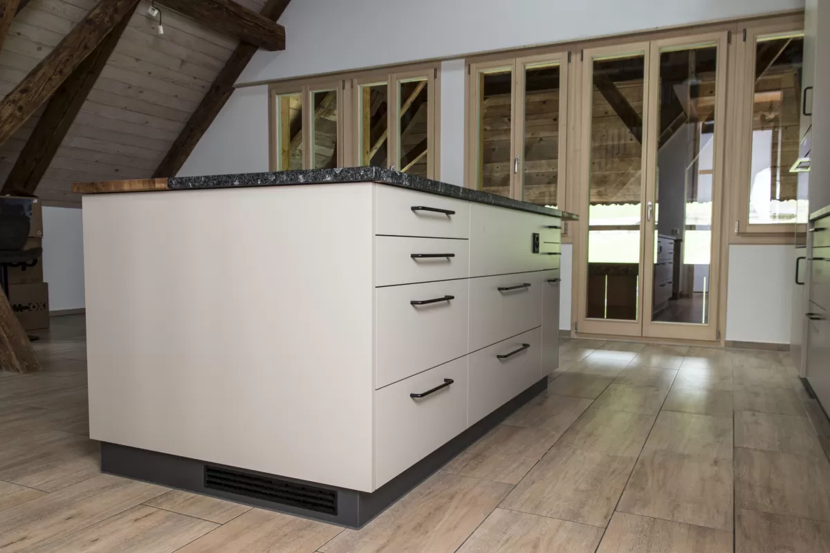 Moderne Küche mit heller Einbauküche, Kochinsel mit Holzarbeitsplatte und integrierten Geräten in einem offenen Wohnraum mit sichtbaren Holzbalken.