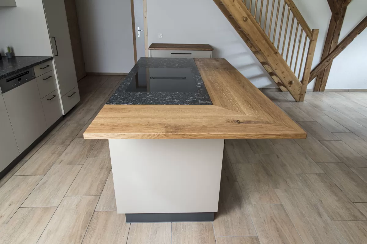 Moderne Küche mit heller Einbauküche, Kochinsel mit Holzarbeitsplatte und integrierten Geräten in einem offenen Wohnraum mit sichtbaren Holzbalken.