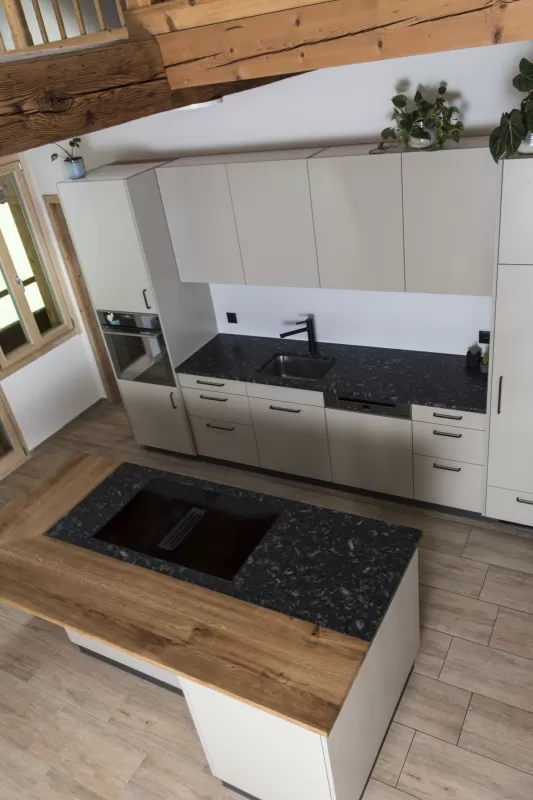 Moderne Küche mit heller Einbauküche, Kochinsel mit Holzarbeitsplatte und integrierten Geräten in einem offenen Wohnraum mit sichtbaren Holzbalken.