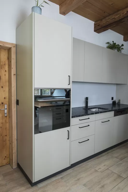 Moderne Küche mit heller Einbauküche, Kochinsel mit Holzarbeitsplatte und integrierten Geräten in einem offenen Wohnraum mit sichtbaren Holzbalken.