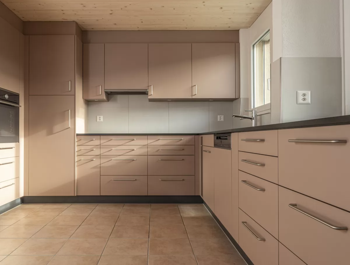 Moderne Küche mit matten, roséfarbenen Fronten, Edelstahlgriffen und hellbraunem Fliesenboden. Die Küche hat eine L-Form, eine dunkle Arbeitsplatte und eine Holzdecke mit natürlicher Maserung.