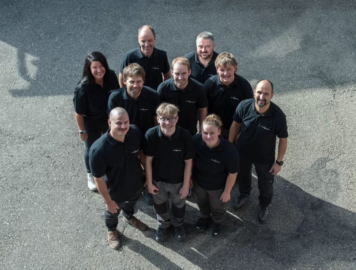 Gruppenfoto des Teams der Zimmerei Hirschi AG: Mitarbeitende in einheitlichen schwarzen Poloshirts stehen im Freien zusammen und blicken lächelnd in die Kamera. Das Bild vermittelt Teamgeist, Zusammenhalt und die persönliche Handschrift des Unternehmens.