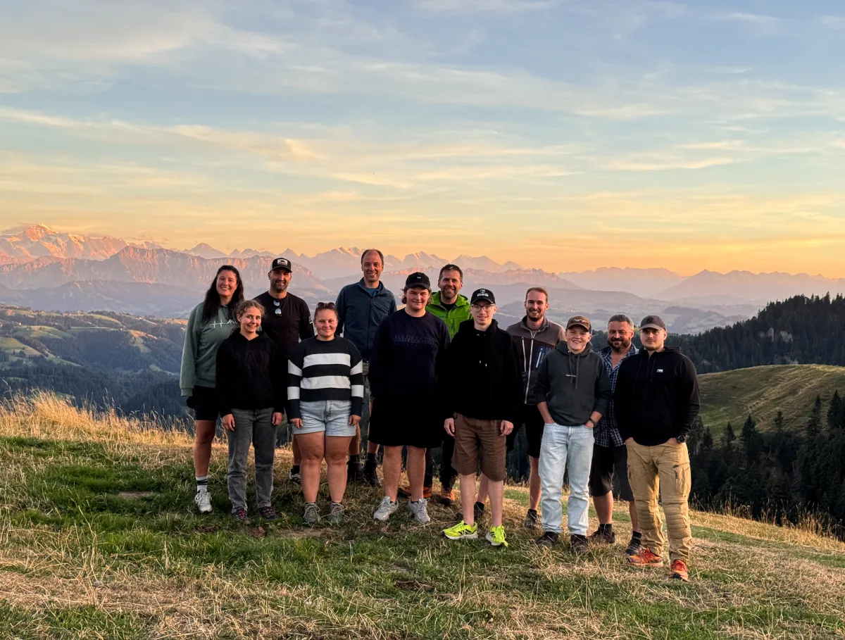 Eine Gruppe von elf Personen steht gemeinsam auf einer grünen Bergwiese und blickt lächelnd in die Kamera. Sie tragen legere Freizeit- und Wanderkleidung. Im Hintergrund erstreckt sich ein weitläufiges Alpenpanorama mit zahlreichen Berggipfeln und bewaldeten Hängen. Die tief stehende Sonne taucht die Landschaft und den Himmel in warme, goldene Farbtöne
