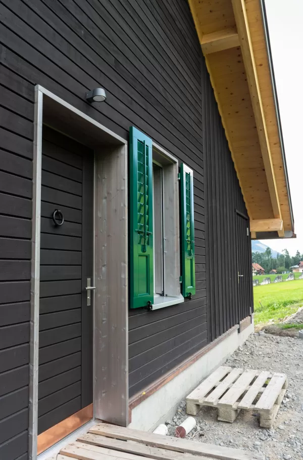 Aussenansicht eines Holzhauses mit dunkel lasierter Fassade, Eingangstür und Fenster mit grünen Holzläden unter einem grossen Dachvorsprung
