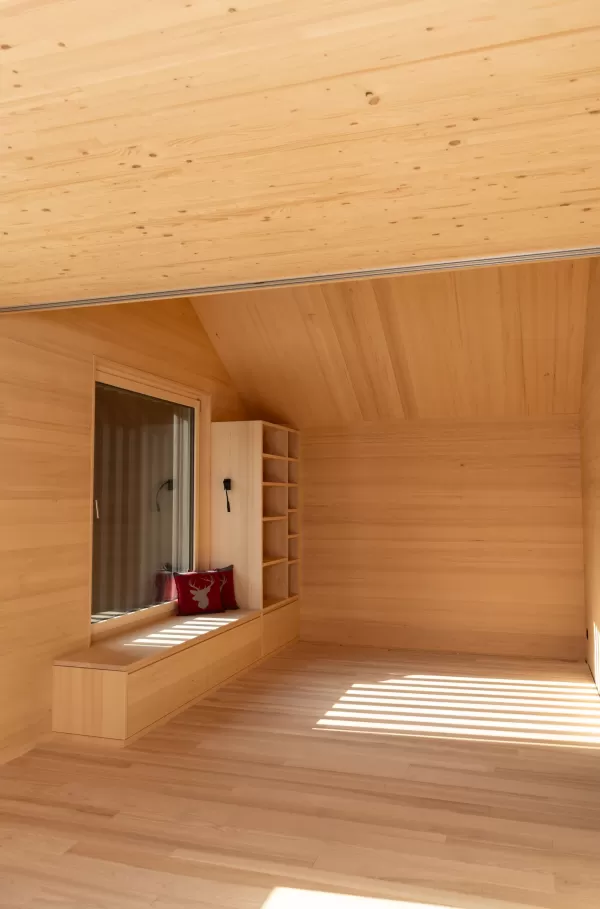 Holzverkleidete Sitznische am Fenster mit integrierten Schubladen, offenen Regalen und roten Zierkissen in einem hellen Innenraum.