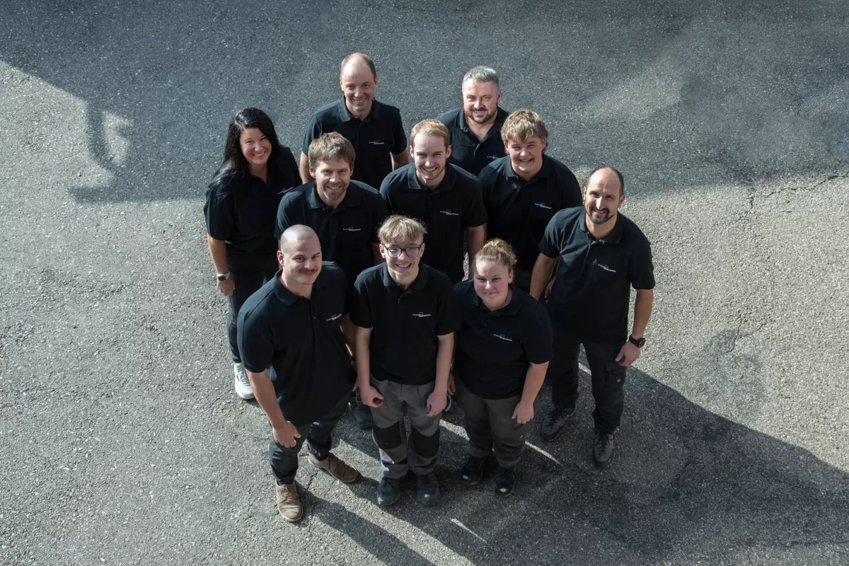 Gruppenfoto des Teams der Zimmerei Hirschi AG: Mitarbeitende in einheitlichen schwarzen Poloshirts stehen im Freien zusammen und blicken lächelnd in die Kamera. Das Bild vermittelt Teamgeist, Zusammenhalt und die persönliche Handschrift des Unternehmens.