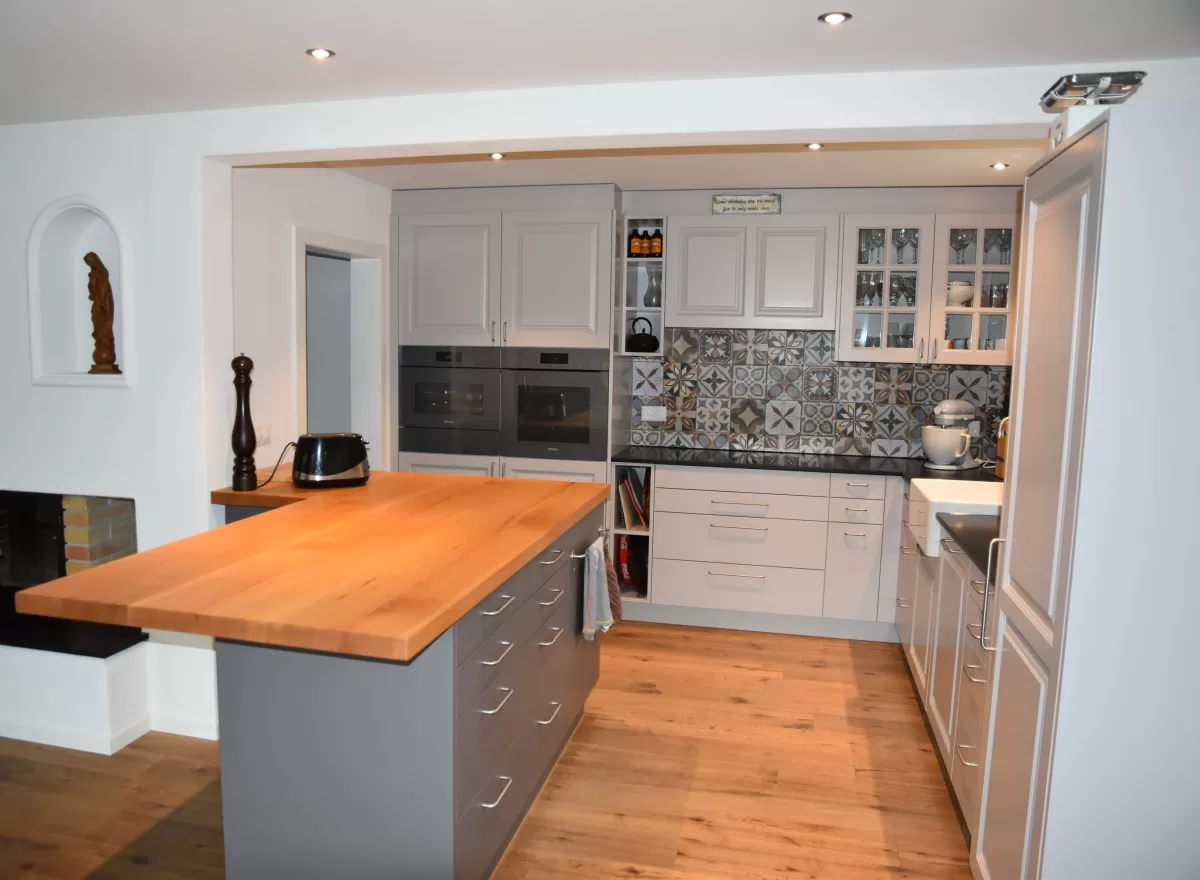 Individuell gestaltete Küche mit hellen Fronten, integrierten Einbaugeräten und grosszügiger Kochinsel mit massiver Holzarbeitsplatte. Der dekorative Fliesenspiegel setzt einen farbigen Akzent und bildet einen Kontrast zu den klaren Linien der Möblierung. Warme Holzböden und die offene Raumgestaltung schaffen eine einladende und wohnliche Atmosphäre.