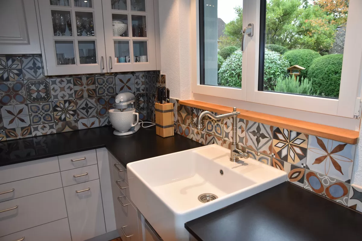 Individuell gestaltete Küche mit hellen Fronten, integrierten Einbaugeräten und grosszügiger Kochinsel mit massiver Holzarbeitsplatte. Der dekorative Fliesenspiegel setzt einen farbigen Akzent und bildet einen Kontrast zu den klaren Linien der Möblierung. Warme Holzböden und die offene Raumgestaltung schaffen eine einladende und wohnliche Atmosphäre.