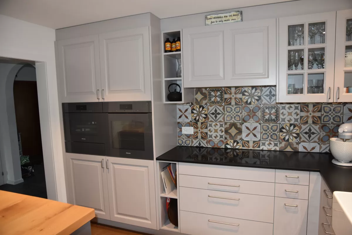 Individuell gestaltete Küche mit hellen Fronten, integrierten Einbaugeräten und grosszügiger Kochinsel mit massiver Holzarbeitsplatte. Der dekorative Fliesenspiegel setzt einen farbigen Akzent und bildet einen Kontrast zu den klaren Linien der Möblierung. Warme Holzböden und die offene Raumgestaltung schaffen eine einladende und wohnliche Atmosphäre.