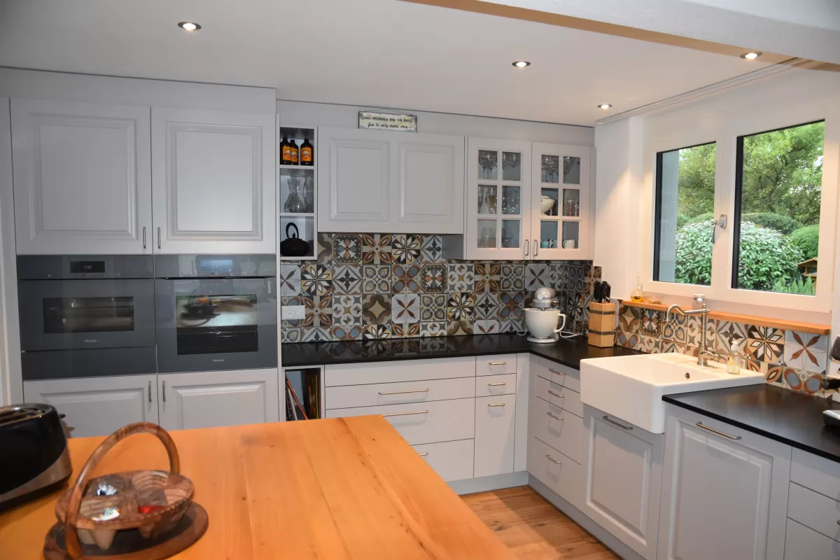 Individuell gestaltete Küche mit hellen Fronten, integrierten Einbaugeräten und grosszügiger Kochinsel mit massiver Holzarbeitsplatte. Der dekorative Fliesenspiegel setzt einen farbigen Akzent und bildet einen Kontrast zu den klaren Linien der Möblierung. Warme Holzböden und die offene Raumgestaltung schaffen eine einladende und wohnliche Atmosphäre.