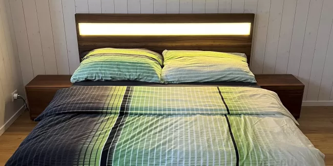 Ein modernes Doppelbett aus dunkel gemasertem Holz steht im Mittelpunkt des Schlafzimmers. Das Kopfteil ist mit einem horizontalen, warm leuchtenden Lichtstreifen versehen, der den Raum sanft ausleuchtet. Auf dem Bett liegt eine Bettwäsche in harmonischen Grün-, Blau- und Weißtönen mit einem feinen Linienmuster. Links und rechts befinden sich passende Nachttische aus dem gleichen Holz. Der Raum hat einen hellen Holzboden sowie weiss vertäfelte Wände, die eine ruhige, natürliche Atmosphäre schaffen.