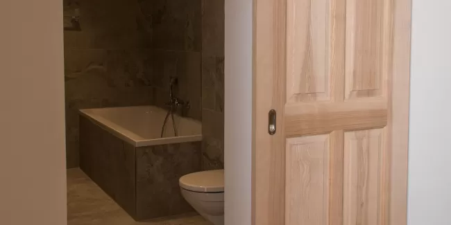Massgefertigte Schiebetür aus Schweizer Eschenholz in heller, natürlicher Optik. Die Tür öffnet den Blick in ein modern gestaltetes Badezimmer mit grossformatigen Steinplatten, einer integrierten Badewanne und einer zeitlosen, schlichten Ausstattung. Die Kombination aus warmem Holz und reduzierten Materialien schafft eine ruhige, hochwertige Wohnatmosphäre.