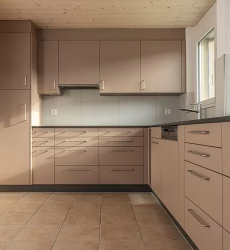 Moderne Küche mit matten, roséfarbenen Fronten, Edelstahlgriffen und hellbraunem Fliesenboden. Die Küche hat eine L-Form, eine dunkle Arbeitsplatte und eine Holzdecke mit natürlicher Maserung.